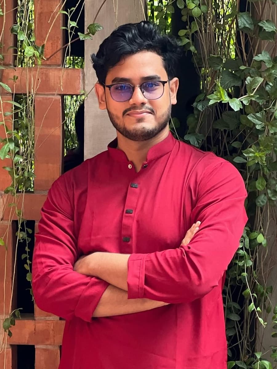 Ishrak Hamim Mahi, arms crossed, in a red panjabi in front of a brick wall with vines